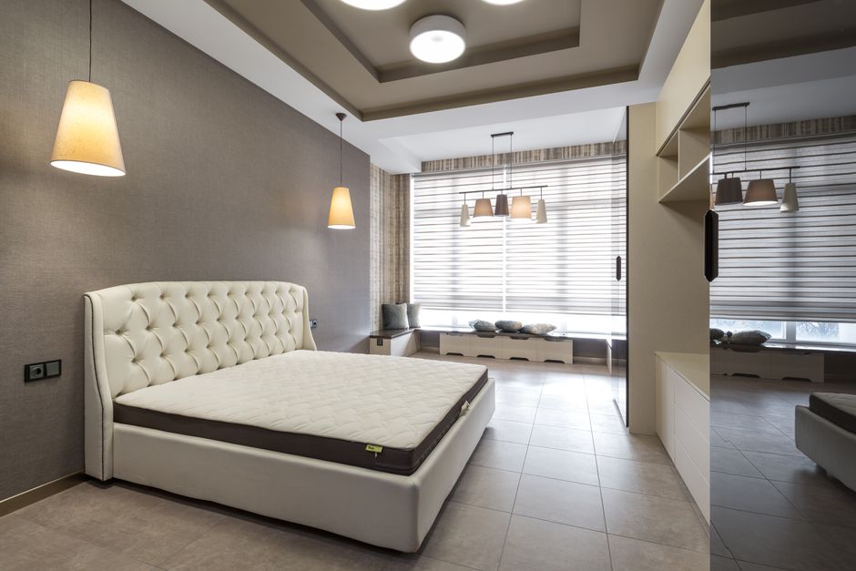 White bed with mattress placed at gray wall against cupboards in spacious bedroom with window and glowing lamps in apartment