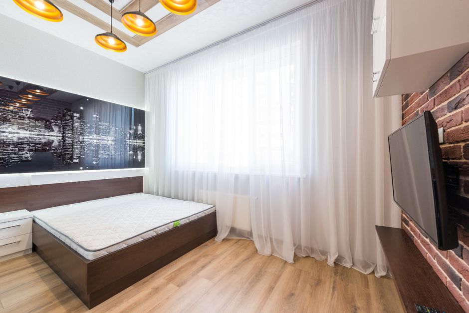 Bed with mattress placed at wall with decorative paintings against TV in modern bedroom with tulle on window in apartment